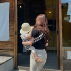 Femme portant un enfant en porte-bébé noir et blanc à motif, devant une maison en bois, en journée ensoleillée